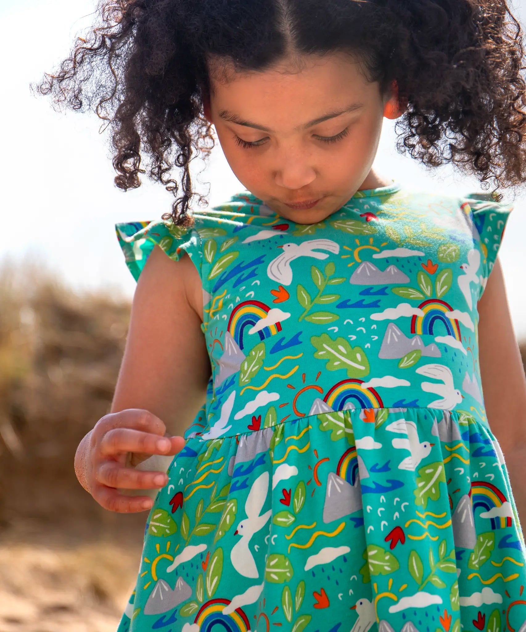 Child wearing Frugi's skater dress in aqua with a repeating seagull and mountain design and frilled shoulders