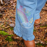 Frugi grace denim wide leg trousers with rainbow and happy embroidery worn by child outside with brown leather boots.