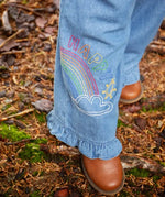 Frugi grace denim wide leg trousers with rainbow and happy embroidery worn by child outside with brown leather boots.