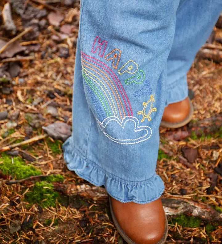 Frugi grace denim wide leg trousers with rainbow and happy embroidery worn by child outside with brown leather boots.