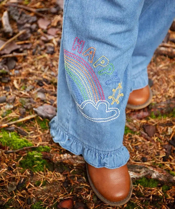 Frugi grace denim wide leg trousers with rainbow and happy embroidery worn by child outside with brown leather boots.