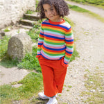A child is stood outside in the sunshine, with their hands in their pockets. The child wears the Frugi Zoe Knitted Jumper - Retro Stripe, which looks very vibrant in colour in the natural day light