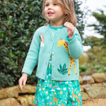 A child outside, wearing the Frugi Colby Cardigan - Moss/Giraffe and a Frugi giraffe dress