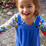 A child outside in the sun, smiling and with their arms out stretched to the side. The child is wearing the Frugi Polly Pinafore Outfit - Blue Tang/Retro Happy. The image showing the adjustable straps with adjust using a button