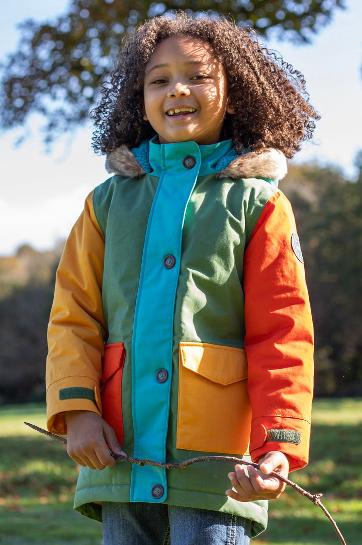Child in a forest wearing Frugi Penhale Parka in Khaki Colourblock - made from PU-coated organic cotton, waterproof 3000HH, to represent Frugi Outerwear available to buy at Babipur.