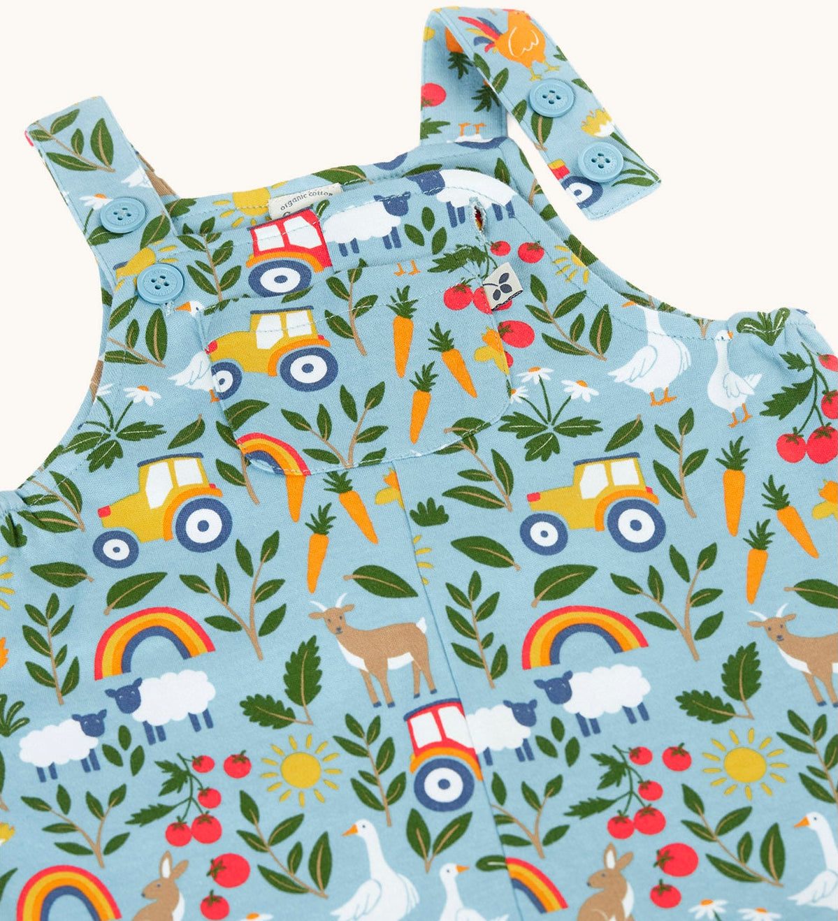 Close up of the light blue Frugi organic cotton dungarees with repeated farm themed print laid flat. Showing 2 poppers open 