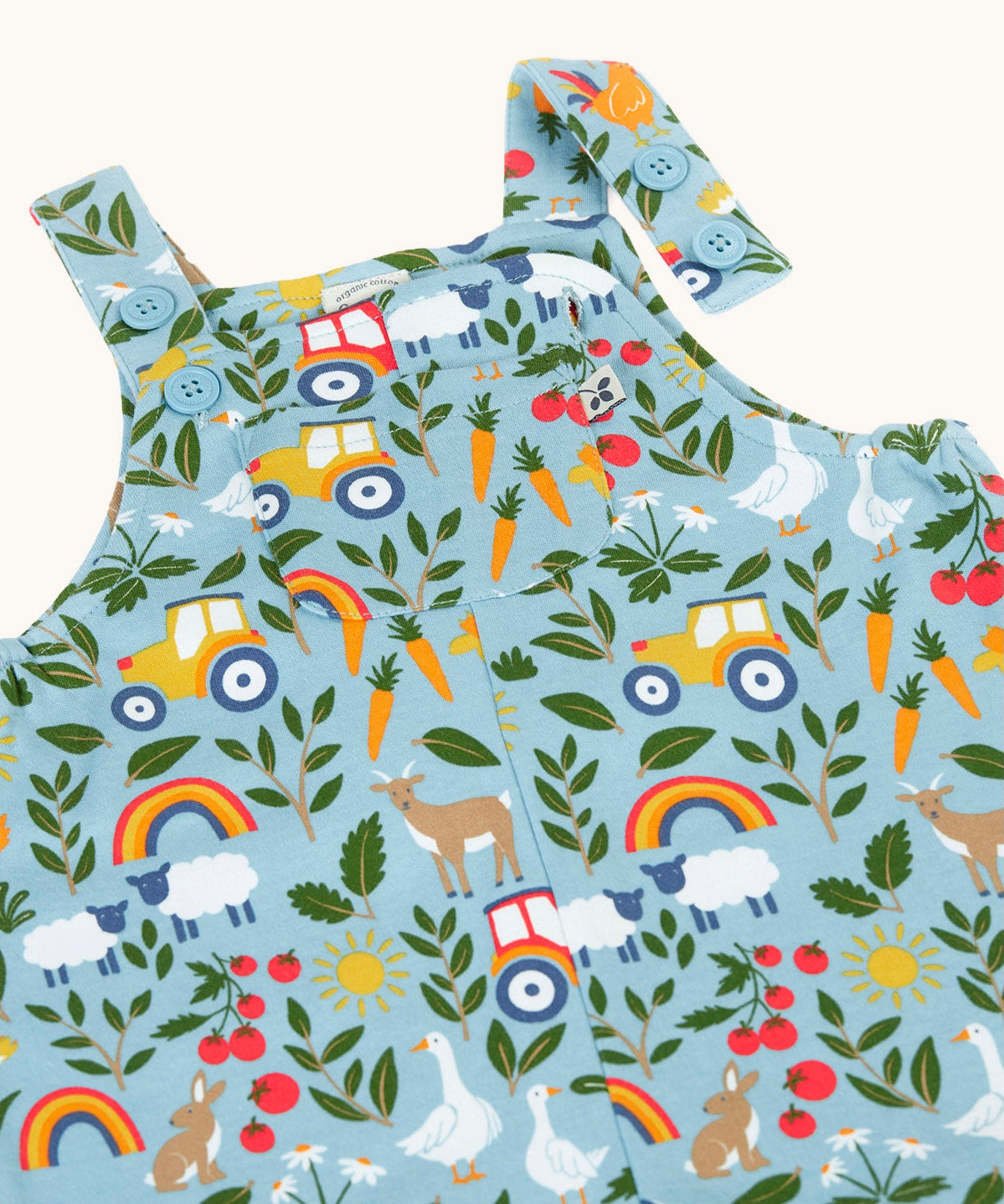 Close up of the light blue Frugi organic cotton dungarees with repeated farm themed print laid flat. Showing 2 poppers open 
