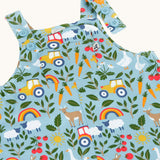 Close up of the light blue Frugi organic cotton dungarees with repeated farm themed print laid flat. Showing 2 poppers open 