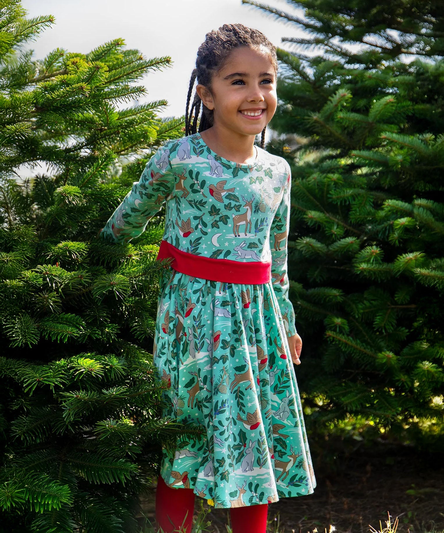 A child standing between two trees and wearing the the Frugi party skater dress in festive forest available at Babipur showing the red tie around the waist