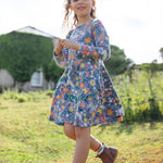 A child happily outside in a grassy green field and a building in the background, wearing the Frugi Sofia Skater Dress - Rainforest Friends