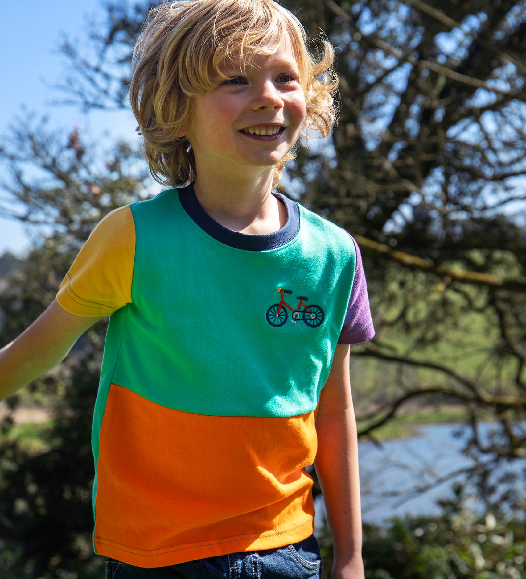 Child wearing Frugi's organic cotton hotchpotch short sleeved kids t-shirt. Shows blue and orange body and purple and yellow sleeves