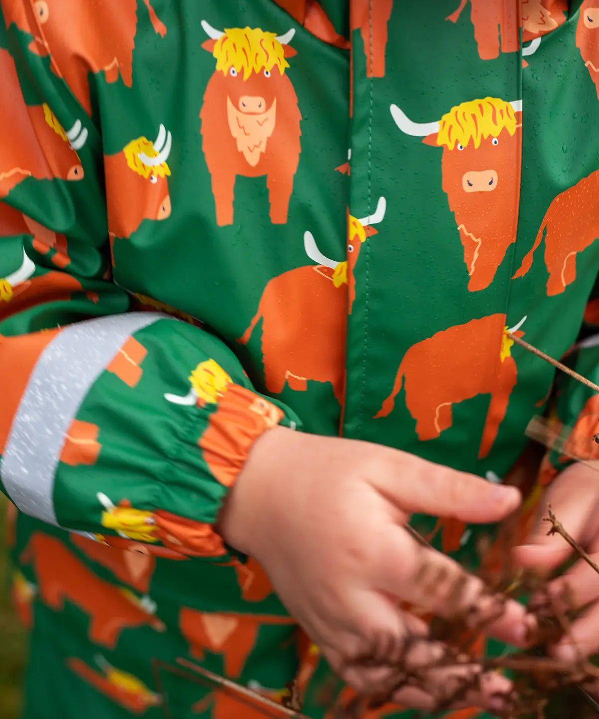 Small child wearing Frugi's waterproof puddle buster coat in highland grazing showing close up of the repeating Highland cow design 