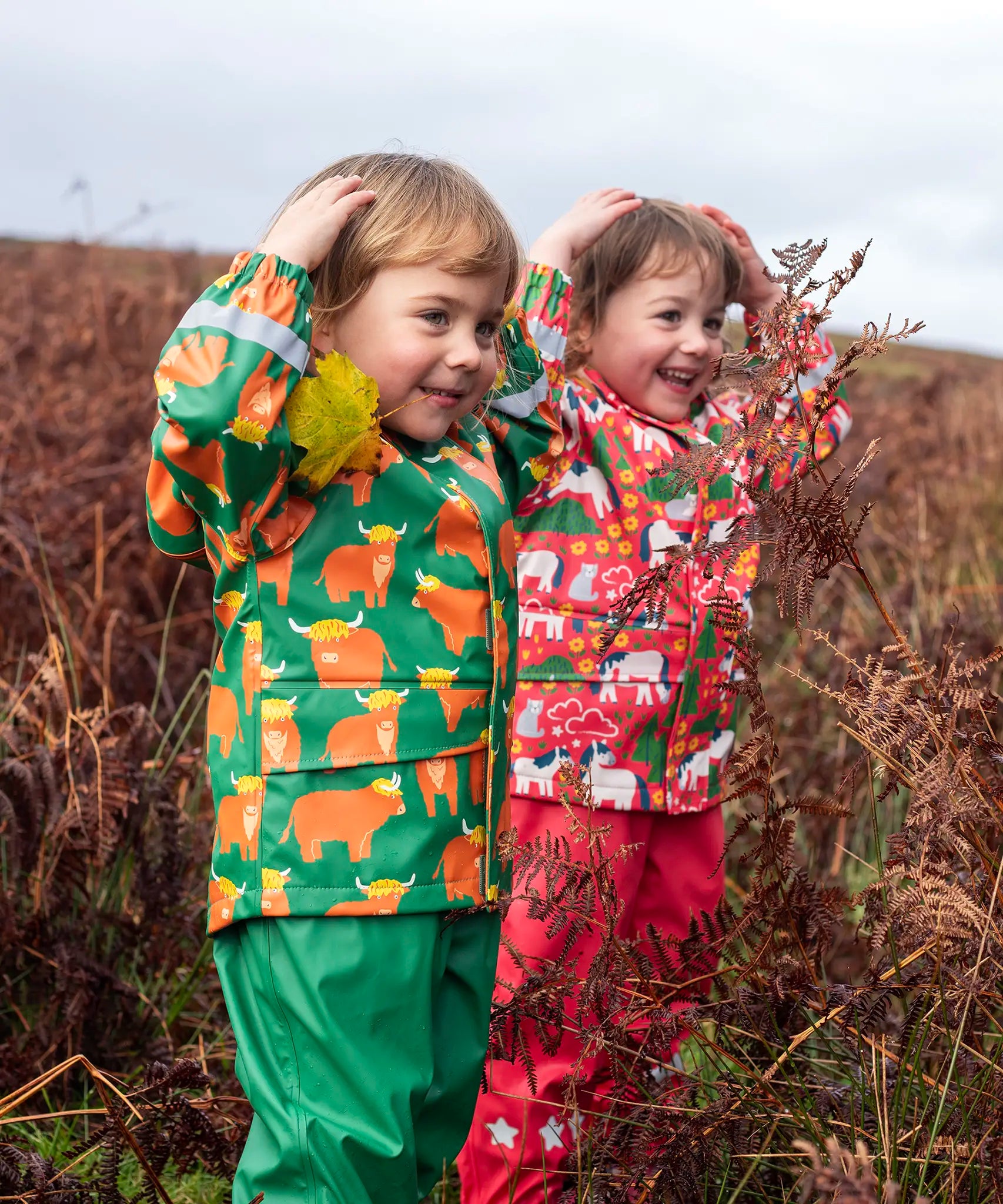 Children wearing Frugi's puddle buster waterproof coats one in green highland grazing design and the other in pink purrfect ponies
