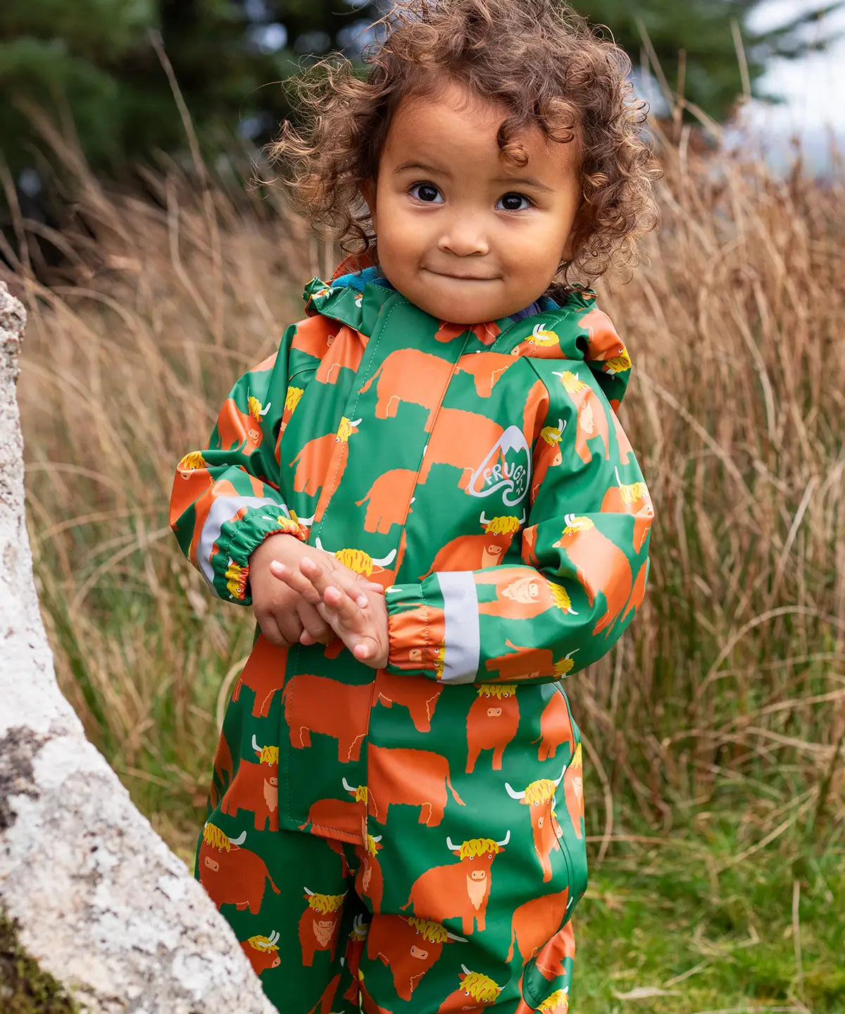 Small child wearing the Frugi waterproof green highland cow puddle buster all in one with the hood down