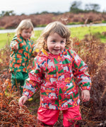 Child wearing Frugi's pink pink puddle buster coat with a repeating horse and tree design in a grassy field.