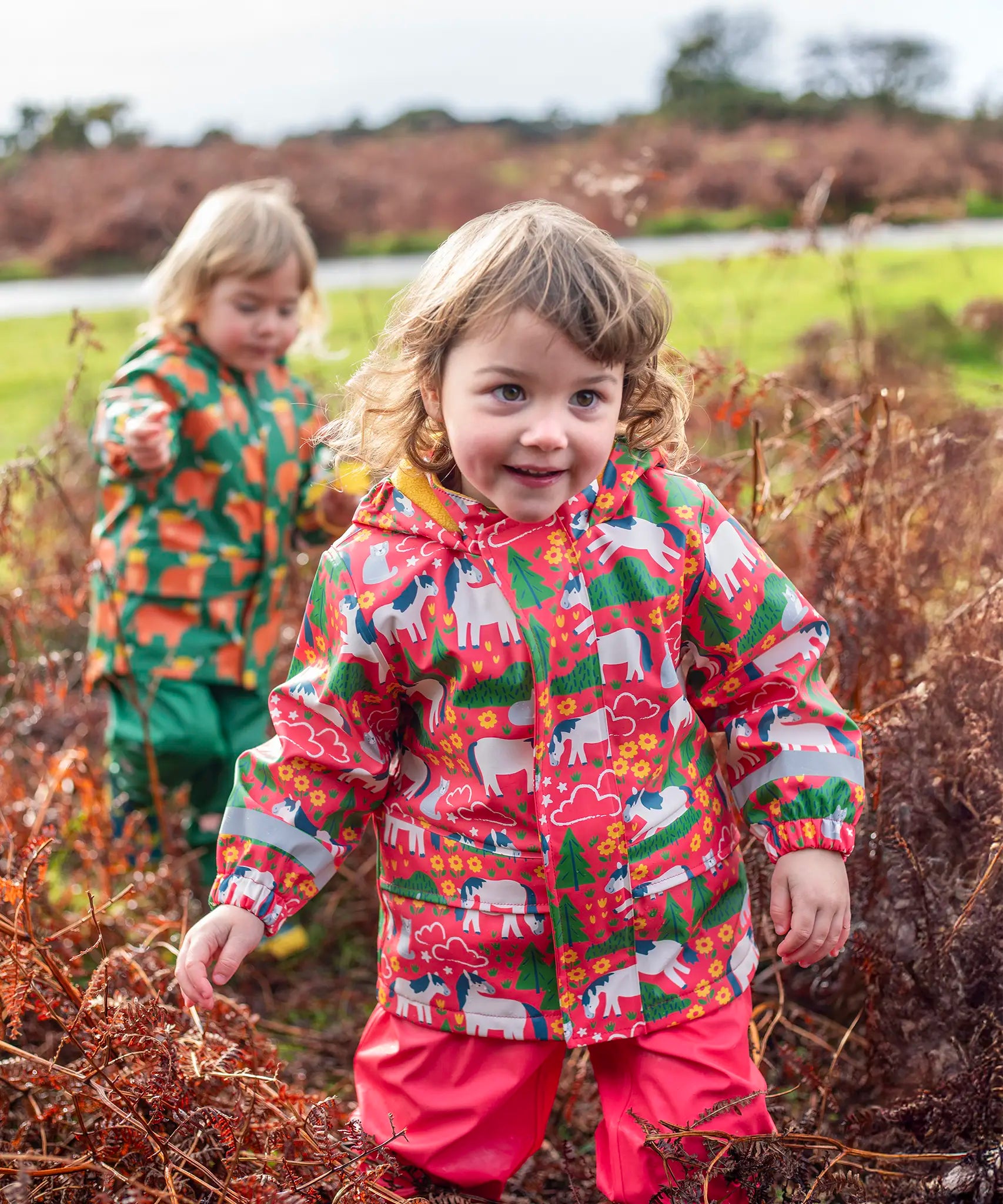 Child wearing Frugi's pink pink puddle buster coat with a repeating horse and tree design in a grassy field.