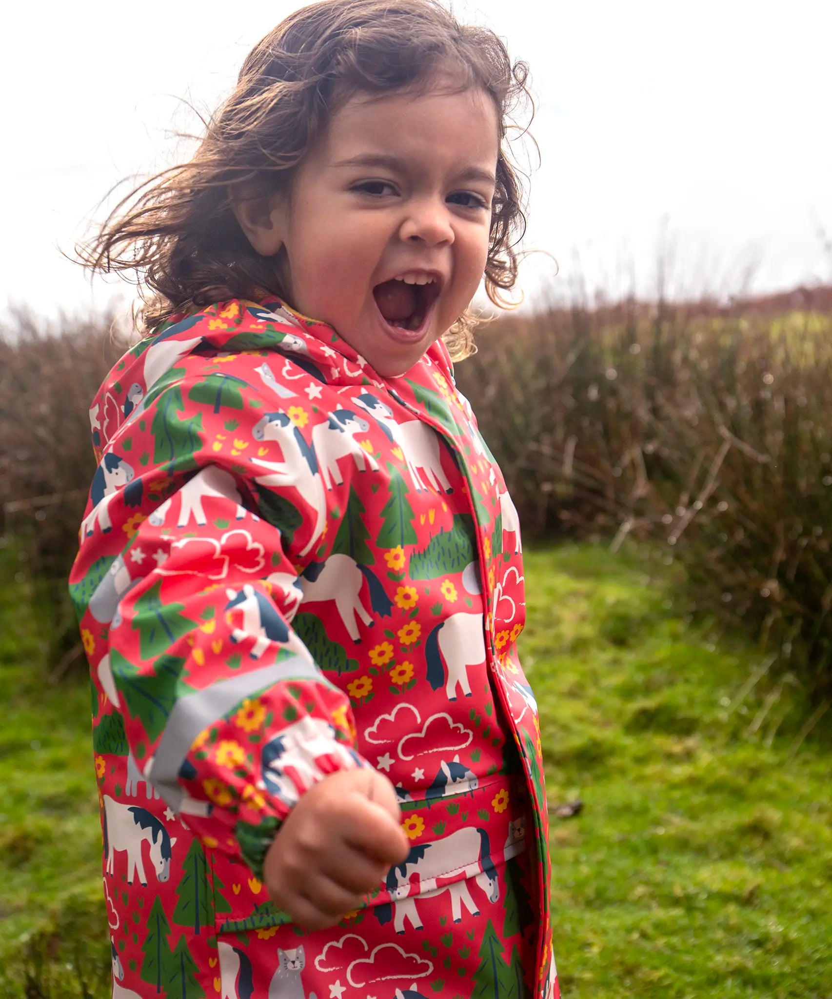Small child wearing Frugi waterproof puddle buster coat in pink purrfect ponies design showing the hood is down 