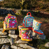 Pair of Frugi purrfect ponies and terrific tractor puddle buster wellington boots standing next to matching little adventure backpacks on a stone wall