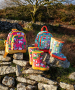 Pair of Frugi purrfect ponies and terrific tractor puddle buster wellington boots standing next to matching little adventure backpacks on a stone wall