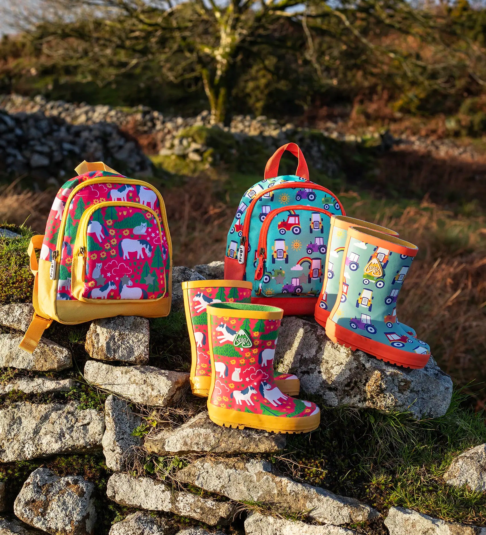 Pair of Frugi purrfect ponies and terrific tractor puddle buster wellington boots standing next to matching little adventure backpacks on a stone wall
