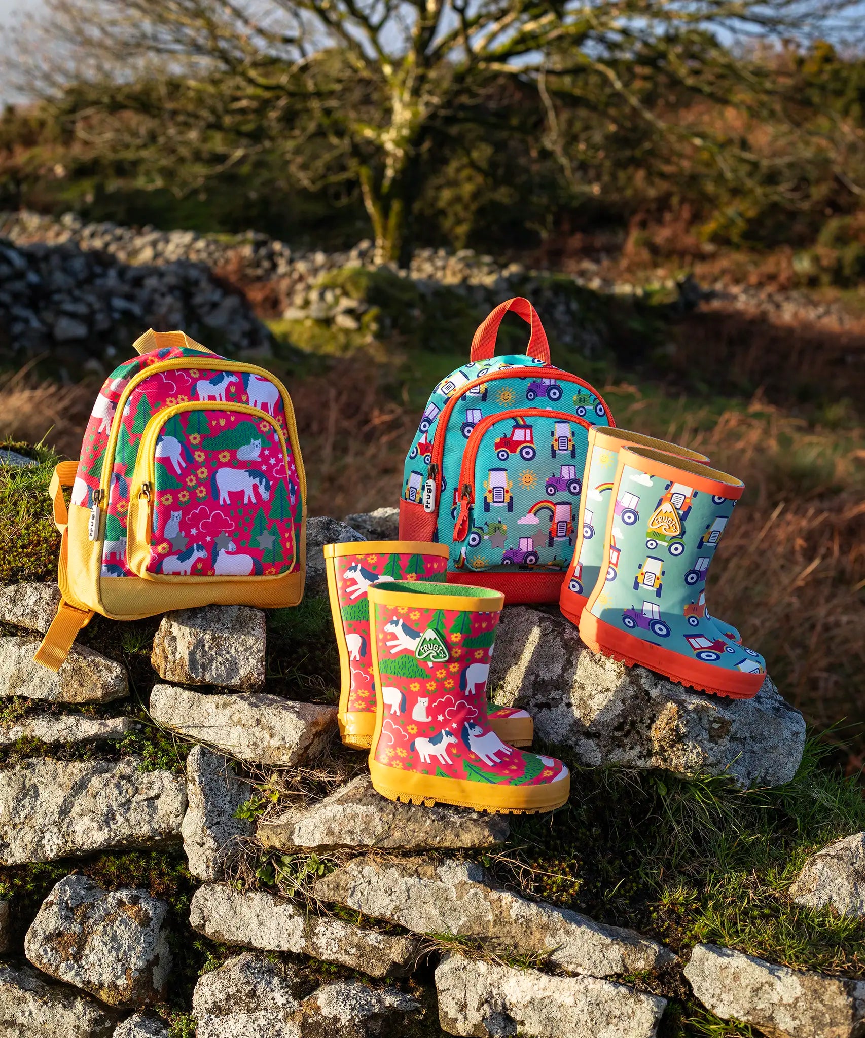 Pair of Frugi purrfect ponies and terrific tractor puddle buster wellington boots standing next to matching little adventure backpacks on a stone wall
