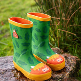 Pair of Frugi natural rubber, waterproof puddle buster green wellingtons with a repeating leaf design and highland cow toe cap standing on a rock 