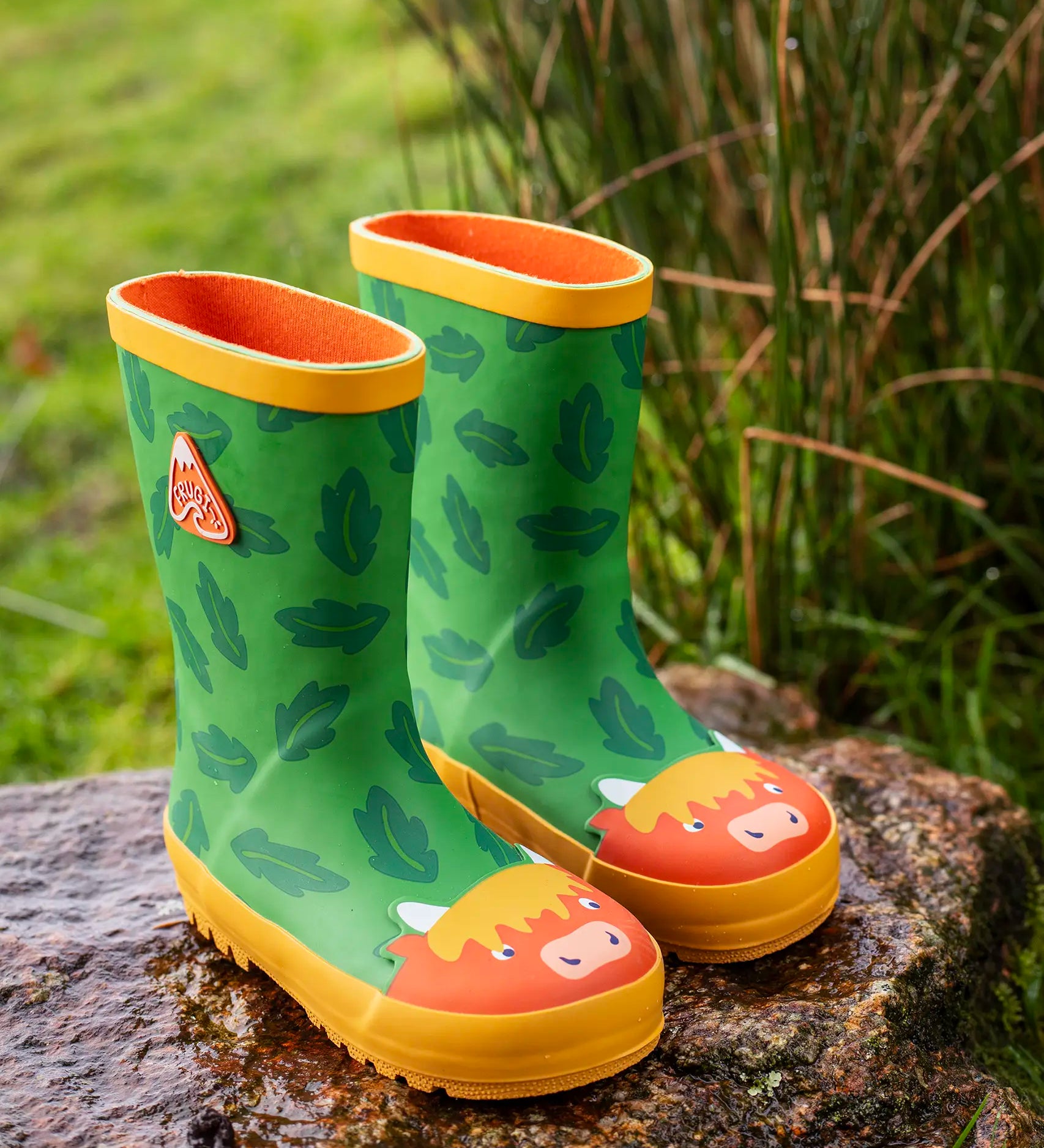 Pair of Frugi natural rubber, waterproof puddle buster green wellingtons with a repeating leaf design and highland cow toe cap standing on a rock 