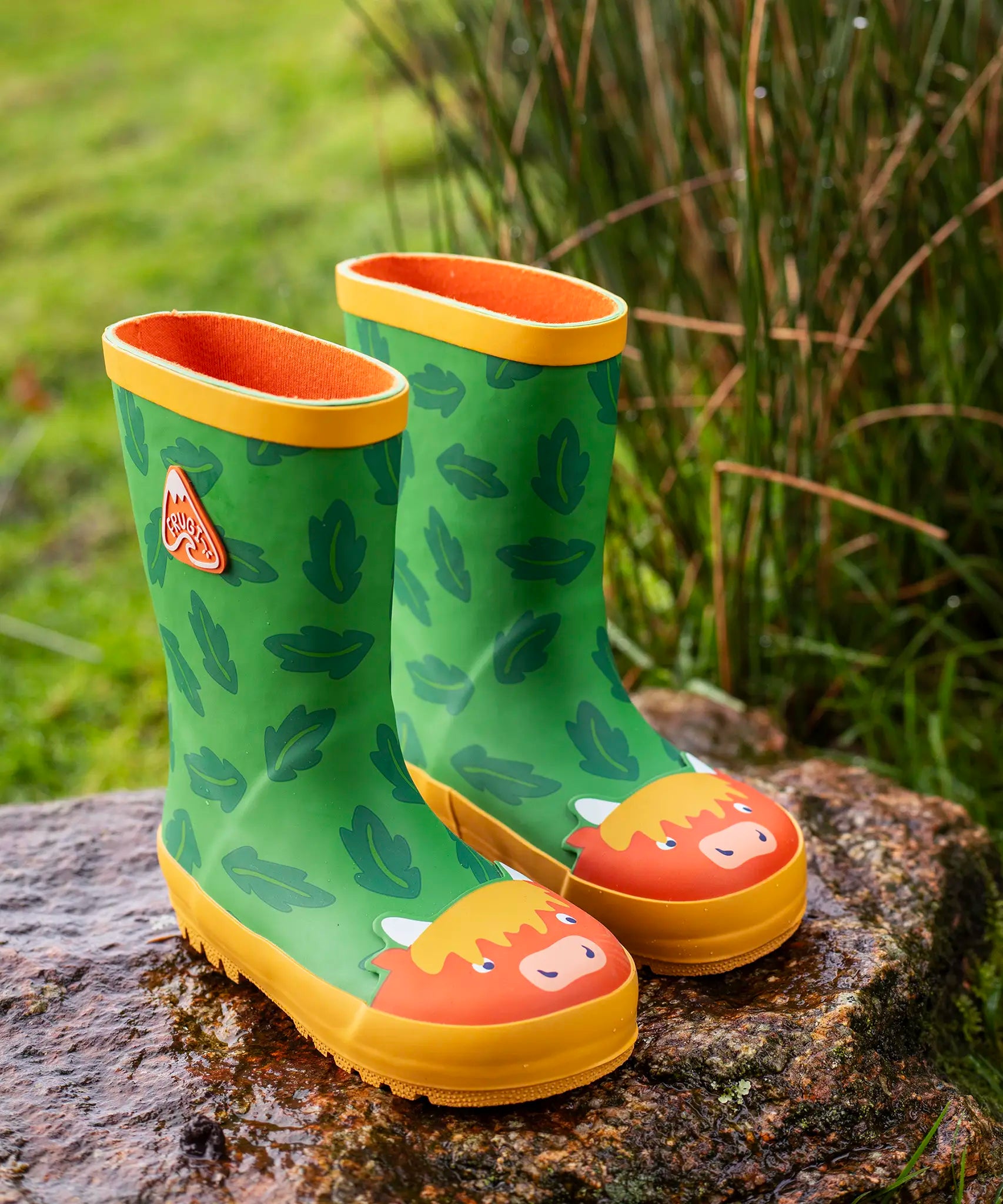 Pair of Frugi natural rubber, waterproof puddle buster green wellingtons with a repeating leaf design and highland cow toe cap standing on a rock 