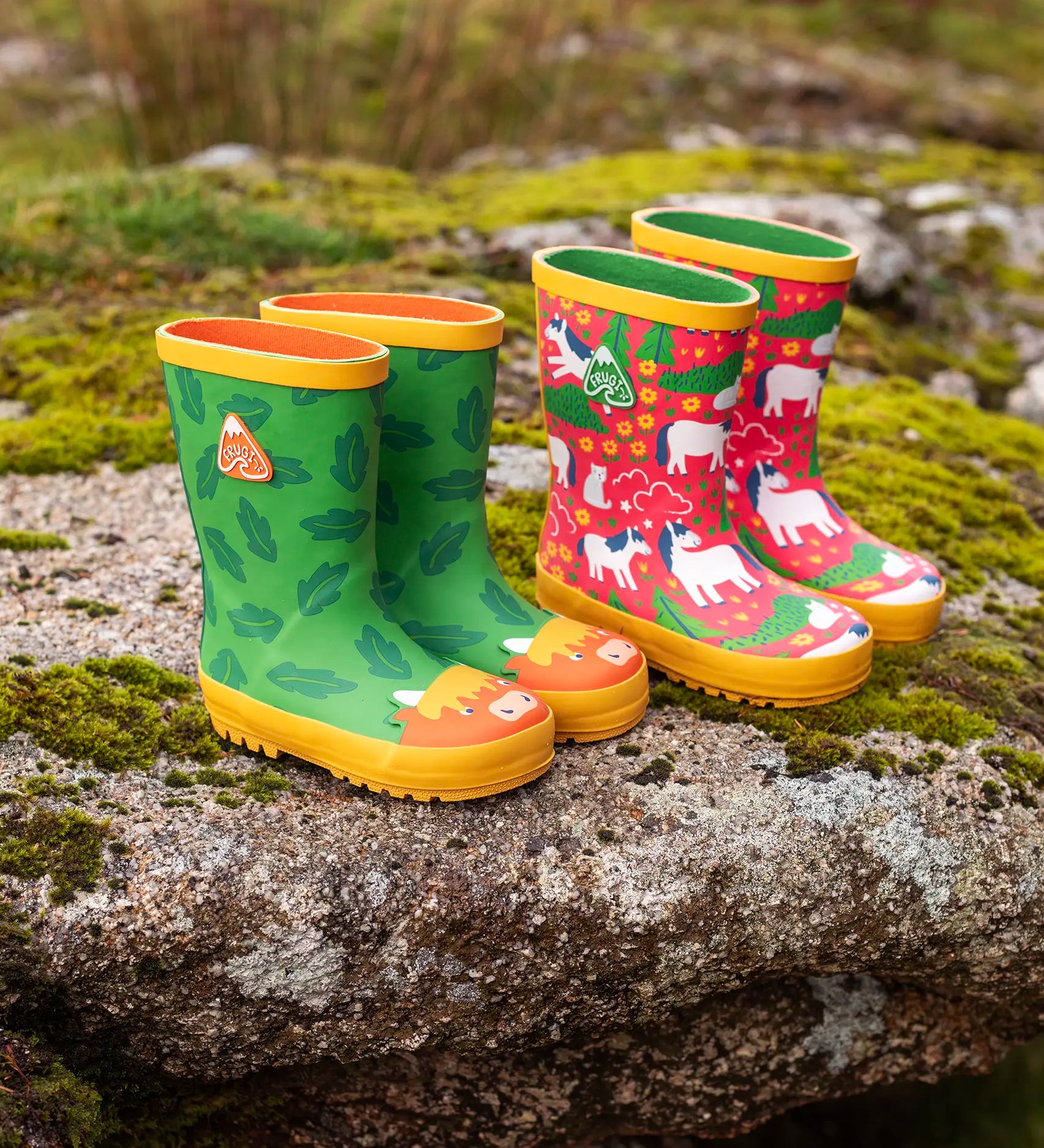 Pair of Frugi natural rubber, waterproof puddle buster wellingtons outdoors showing a green with a repeating leaf design and highland cow toe cap and a pink with repeating white ponies design