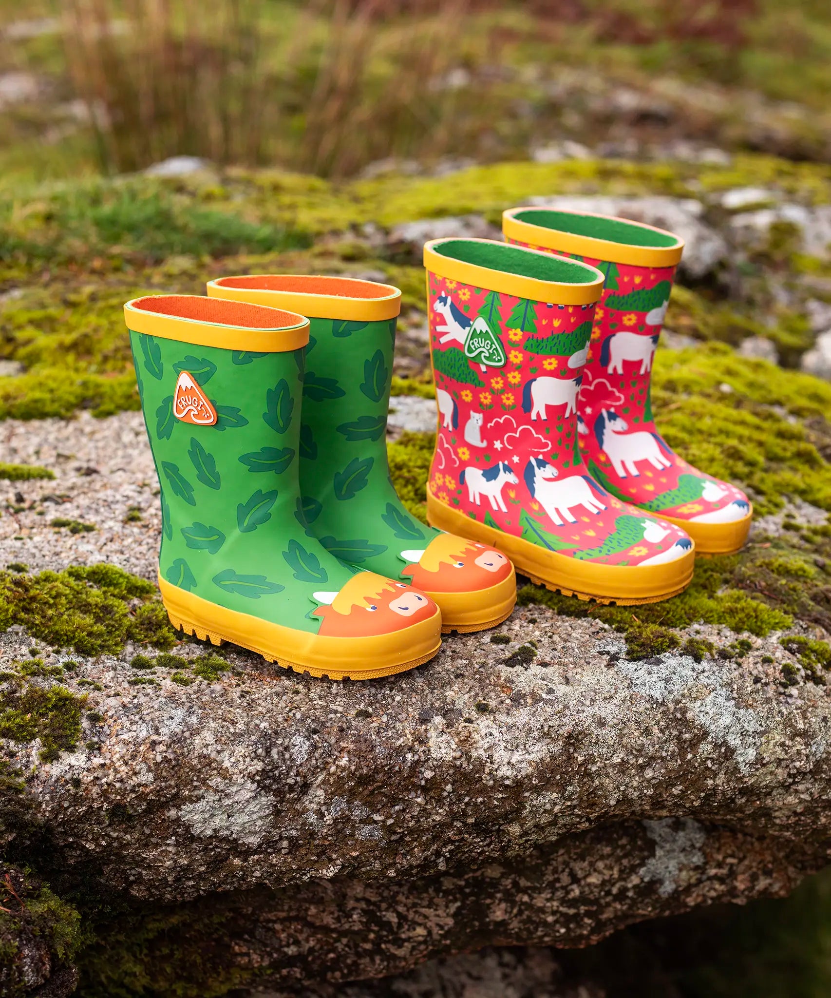 Pair of Frugi natural rubber, waterproof puddle buster wellingtons outdoors showing a green with a repeating leaf design and highland cow toe cap and a pink with repeating white ponies design