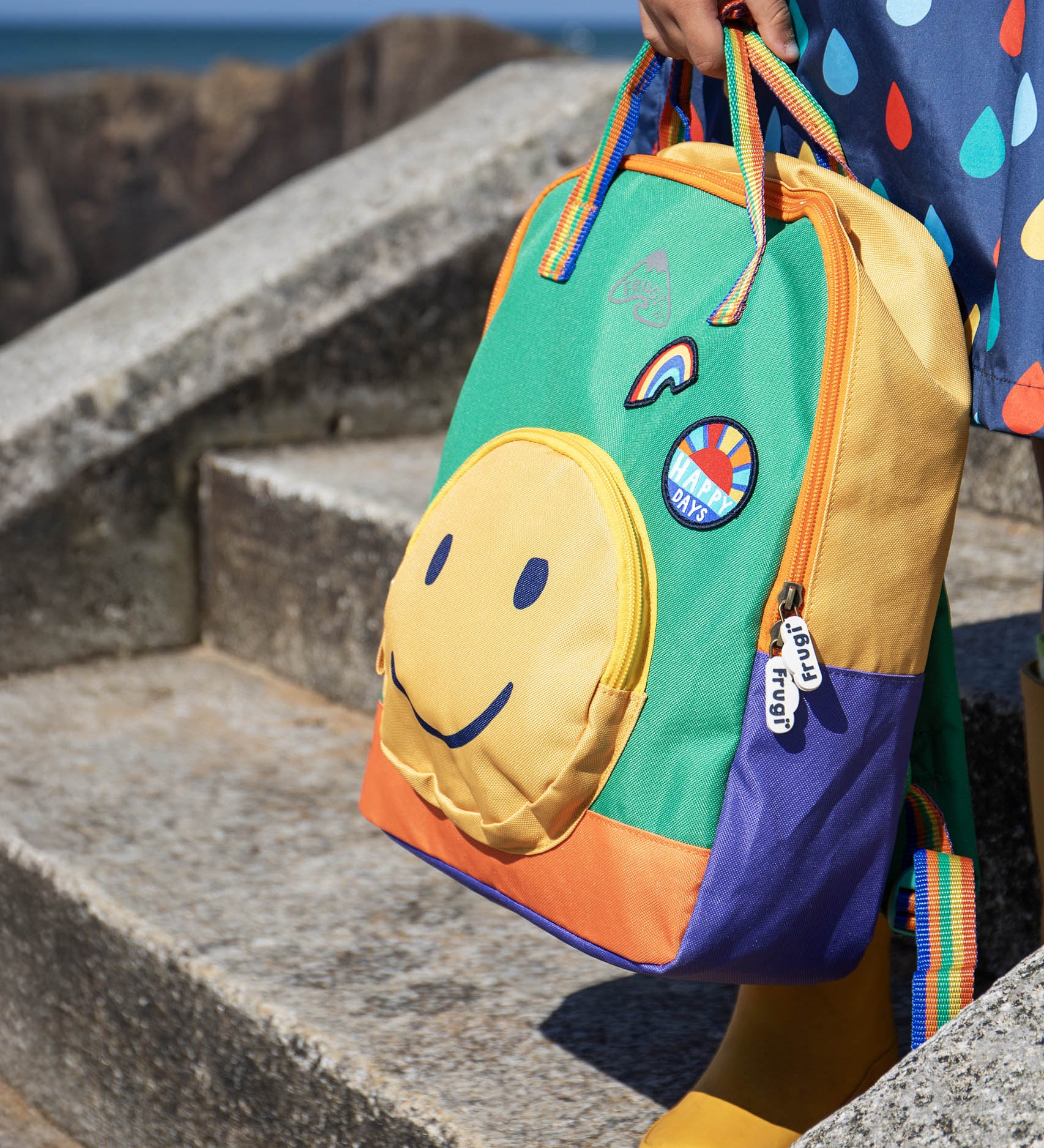 A close up of a child standing on concrete stairs and holding the Frugi green and yellow backpack with a smiling face as a front pocket