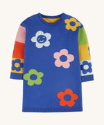 The blue body and colour-block side of the Frugi Zola Reversible Knitted Dress - Blue Tang/Retro Floral. A soft, royal blue, organic cotton knit dress, with colour-block sleeves in yellow, red, green, pink and light blue colour, and colourful bold flower prints 