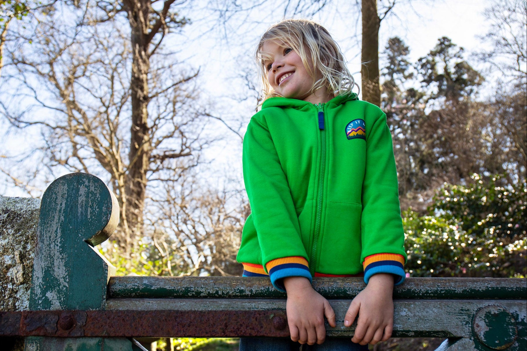 A child standing outside on a gate and wearing the green Jersey style Frugi helston reversible hoodie available at Babipur.