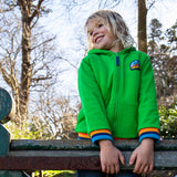 A child standing outside on a gate and wearing the green Jersey style Frugi helston reversible hoodie available at Babipur.