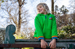 A child standing outside on a gate and wearing the green Jersey style Frugi helston reversible hoodie available at Babipur.
