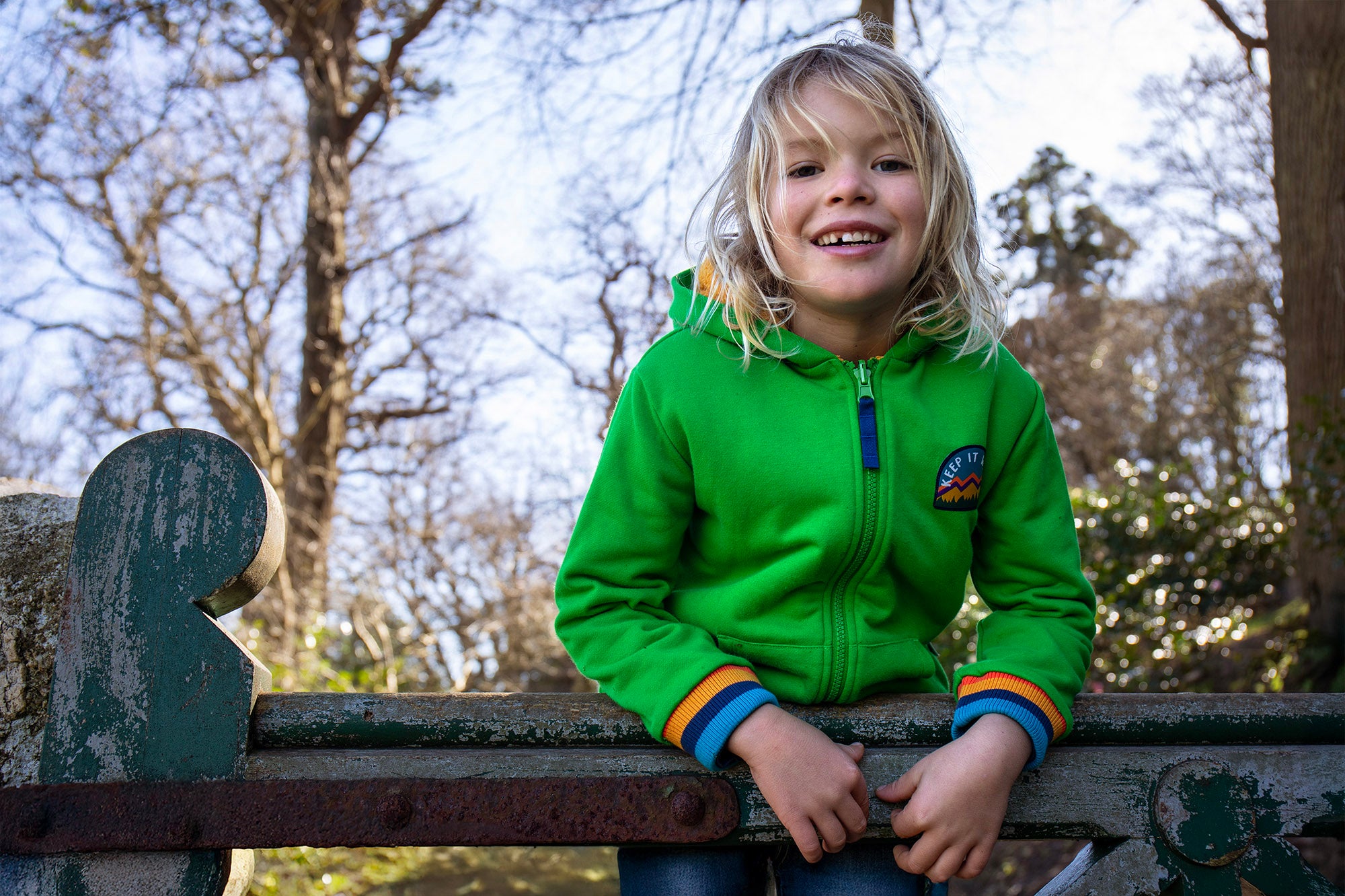 A child standing over a gate and wearing the green Jersey style Frugi helston reversible hoodie available at Babipur.