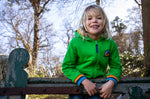A child standing over a gate and wearing the green Jersey style Frugi helston reversible hoodie available at Babipur.