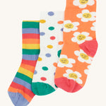 Frugi Rock My Socks - Stay Wild 3 Pack. 3 pairs of soft, organic cotton, kids socks in the Stay Wild prints. from left to right, the left sock is a rainbow stripe print in lilac, orange, red, green and yellow. The middle sock is white with colourful polka dots and a green toe and heel, and the last sock is orange with white, smiling flowers and a red toe and heel.