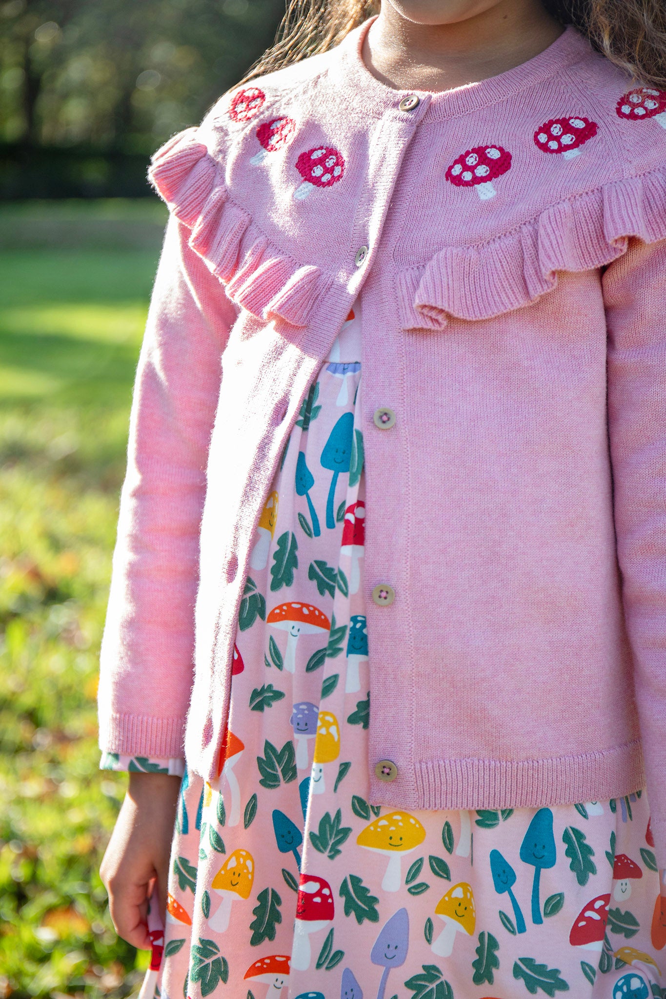 A child wearing the Frugi pink marl Romi cardigan with a matching skater dress available at Babipur. A GOTS organic cotton pink cardigan with a red toadstool embroidery running along the top of the cardigan.