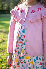 A child wearing the Frugi pink marl Romi cardigan with a matching skater dress available at Babipur. A GOTS organic cotton pink cardigan with a red toadstool embroidery running along the top of the cardigan.