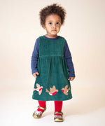 A child wearing the Frugi Rosalie Reversible Baby and Toddler Dress on the green cord side with red tights and gold shoes. One side of this reversible dress features robin redbreast appliques on a Holly green cord with the other side featuring a rainbow coloured check. This dress is a part of a wide range of organic cotton clothing for children available here at Babipur.