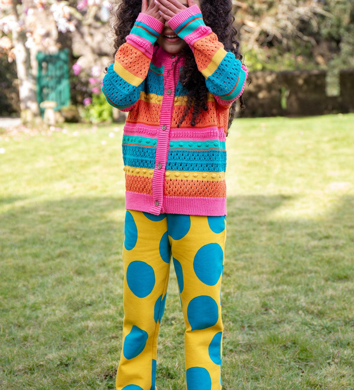 A child standing outside and wearing the Frugi rowe knitted cardigan in rainbow. A bright and bold rainbow striped knitted cardigan with button fasteners available at Babipur.