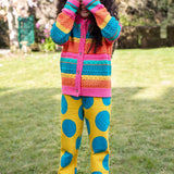 A child standing outside and wearing the Frugi rowe knitted cardigan in rainbow. A bright and bold rainbow striped knitted cardigan with button fasteners available at Babipur.