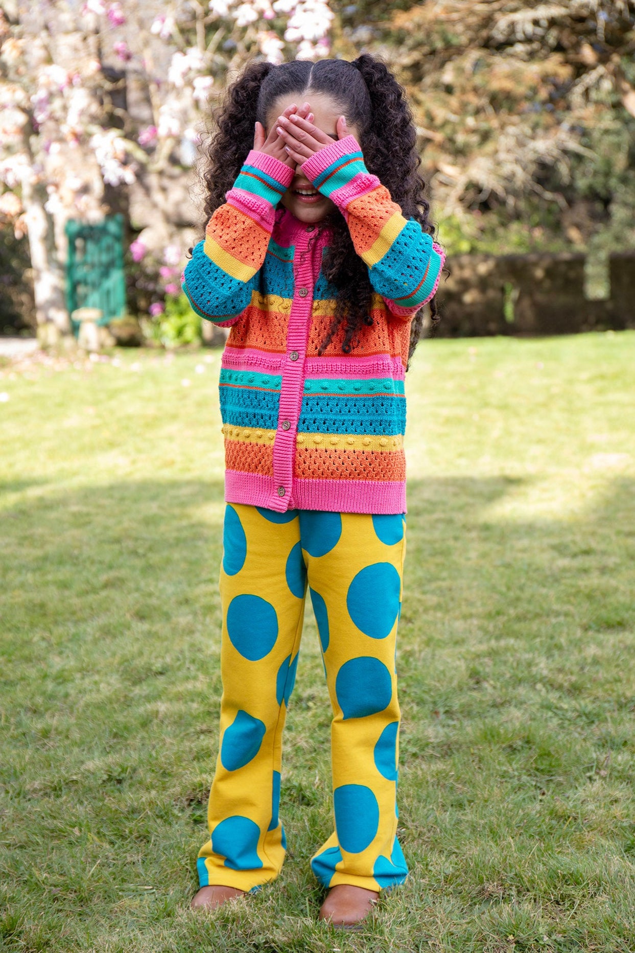 A child standing outside and wearing the Frugi rowe knitted cardigan in rainbow. A bright and bold rainbow striped knitted cardigan with button fasteners available at Babipur.