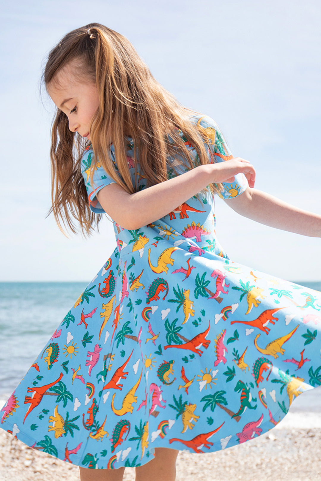 Child on beach wearing Frugi organic cotton twirl dress in blue with colourful dinosaurs. To represent Frugi Spring Sale at babipur
