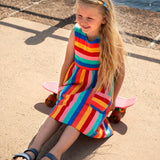 Child wearing Frugi's Samantha rainbow stripe Summer dress and sitting on a small skateboard on the promenade