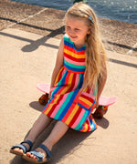 Child wearing Frugi's Samantha rainbow stripe Summer dress and sitting on a small skateboard on the promenade
