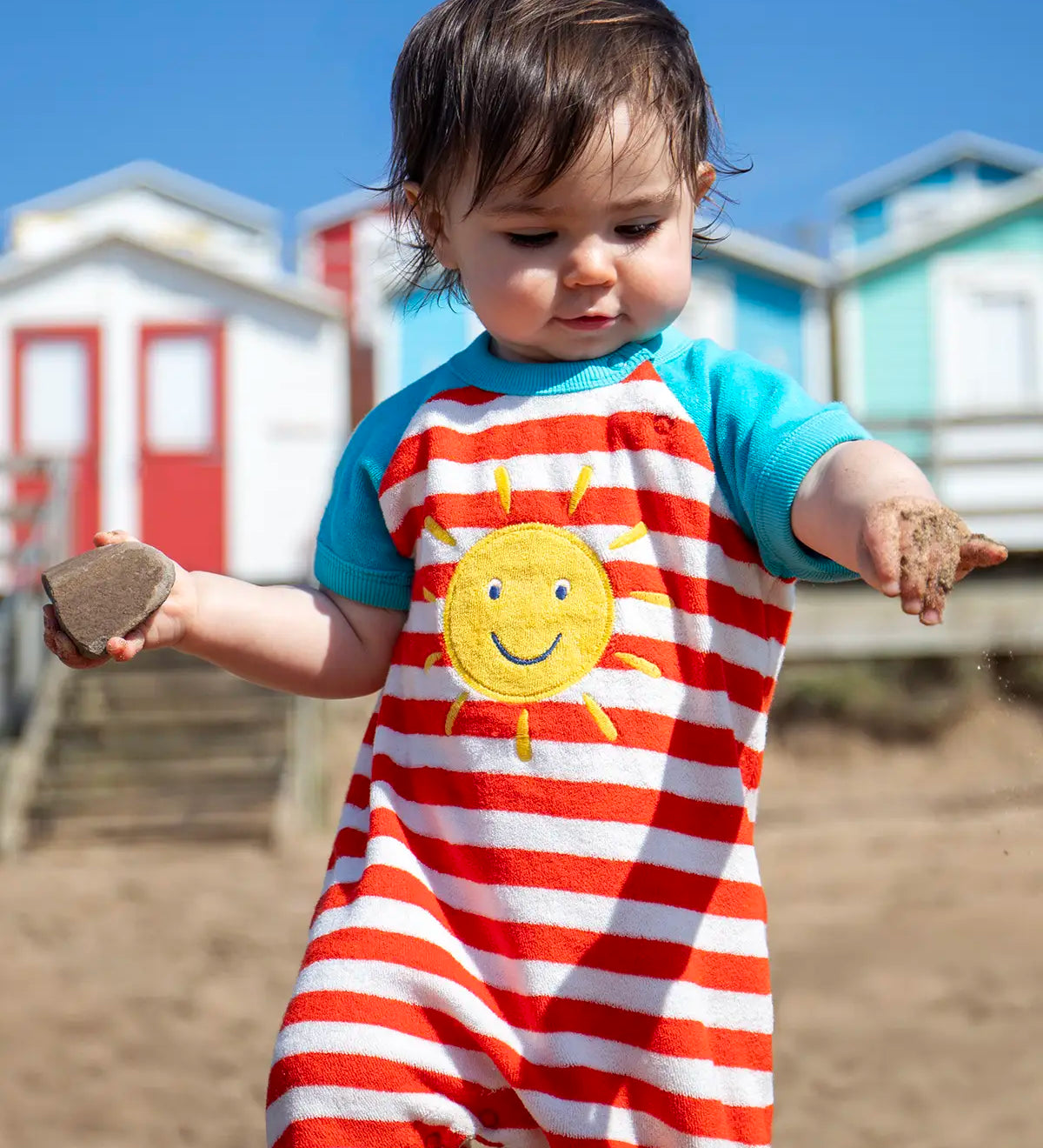 Baby wearing Frugi's towelling romper with blue sleeves, white and red stripe body and yellow sun patch with beach huts in the background