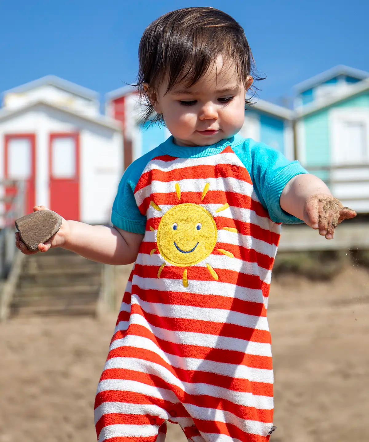 Baby wearing Frugi's towelling romper with blue sleeves, white and red stripe body and yellow sun patch with beach huts in the background