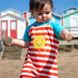 Baby wearing Frugi's towelling romper with blue sleeves, white and red stripe body and yellow sun patch with beach huts in the background